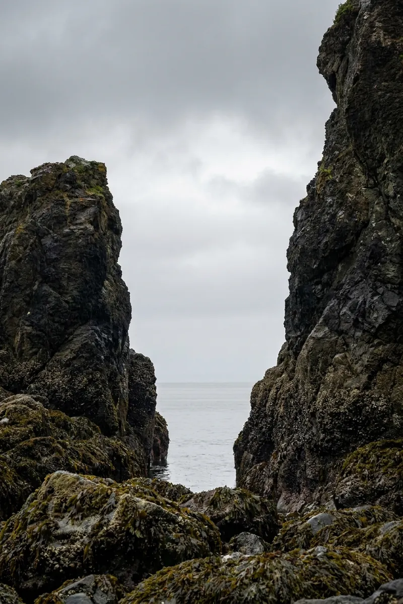 A gap between two dark sea stacks opening onto pale flat ocean and cloud