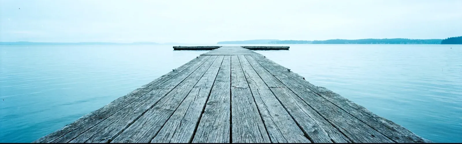 A weathered wooden dock running out into calm Vancouver Island water on a bright morning, a distant shoreline beyond.