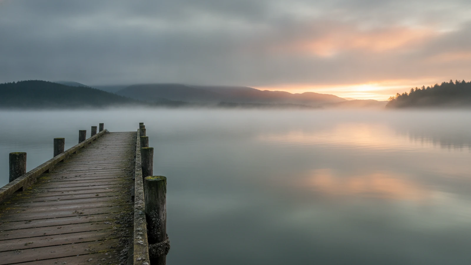 A weathered wooden dock reaching into calm grey-green water at dawn, low mist over the strait, forested hills faint on the far shore.