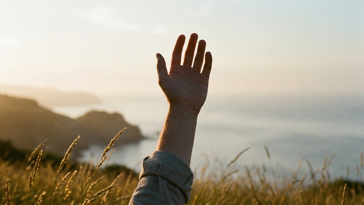A single open hand and forearm raised against a hazy backlit morning sky above coastal grass, a dark headland to the left and pale sea to the right.