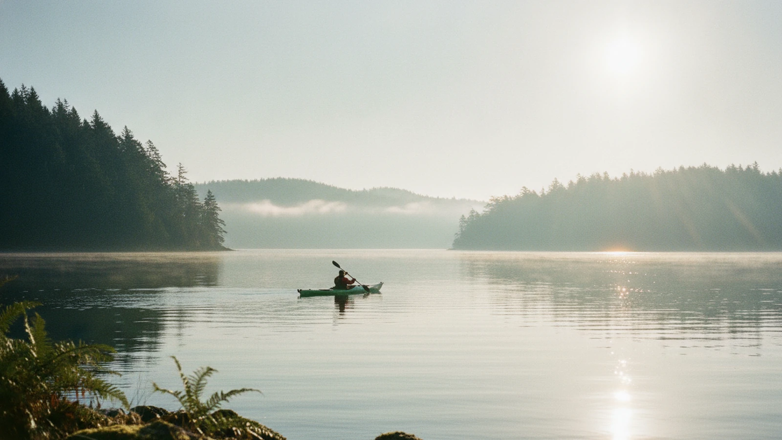 A lone kayaker paddling across a calm, misty bay in low winter sun, fir-covered hills on both shores and a band of sunlight glinting on the water.